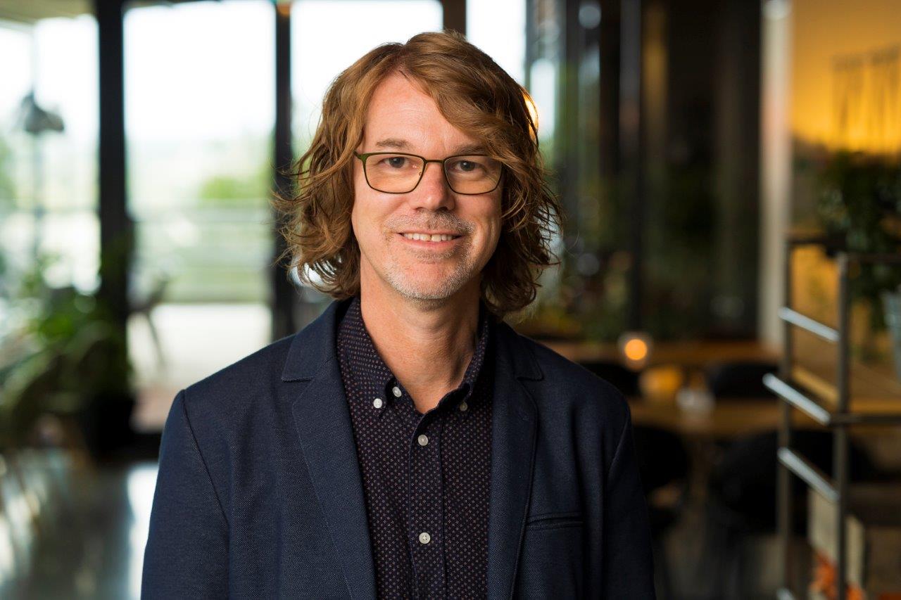 Man med axellångt hår och glasögon i kavaj, ler inomhus i ett ljust, modernt kontorsutrymme.