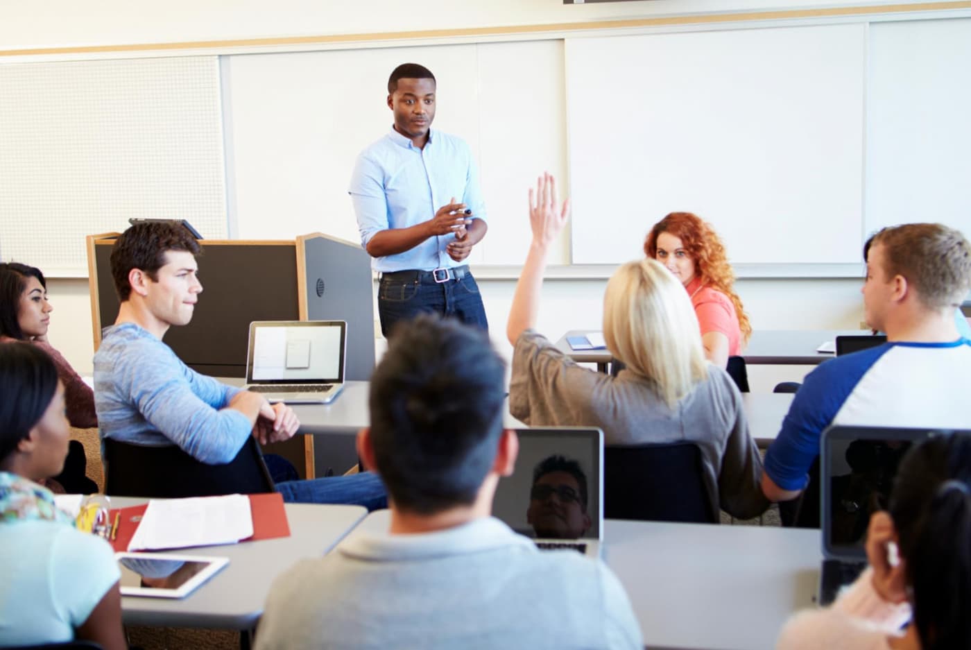 Lärare står längst fram i ett klassrum och undervisar studenter; en student räcker upp handen.